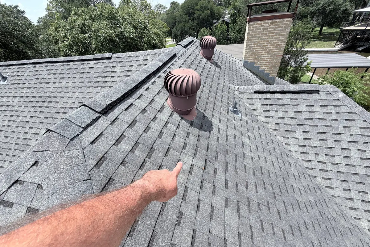 A hand pointing to a shallow dip in a shingle roof near a turbine vent.